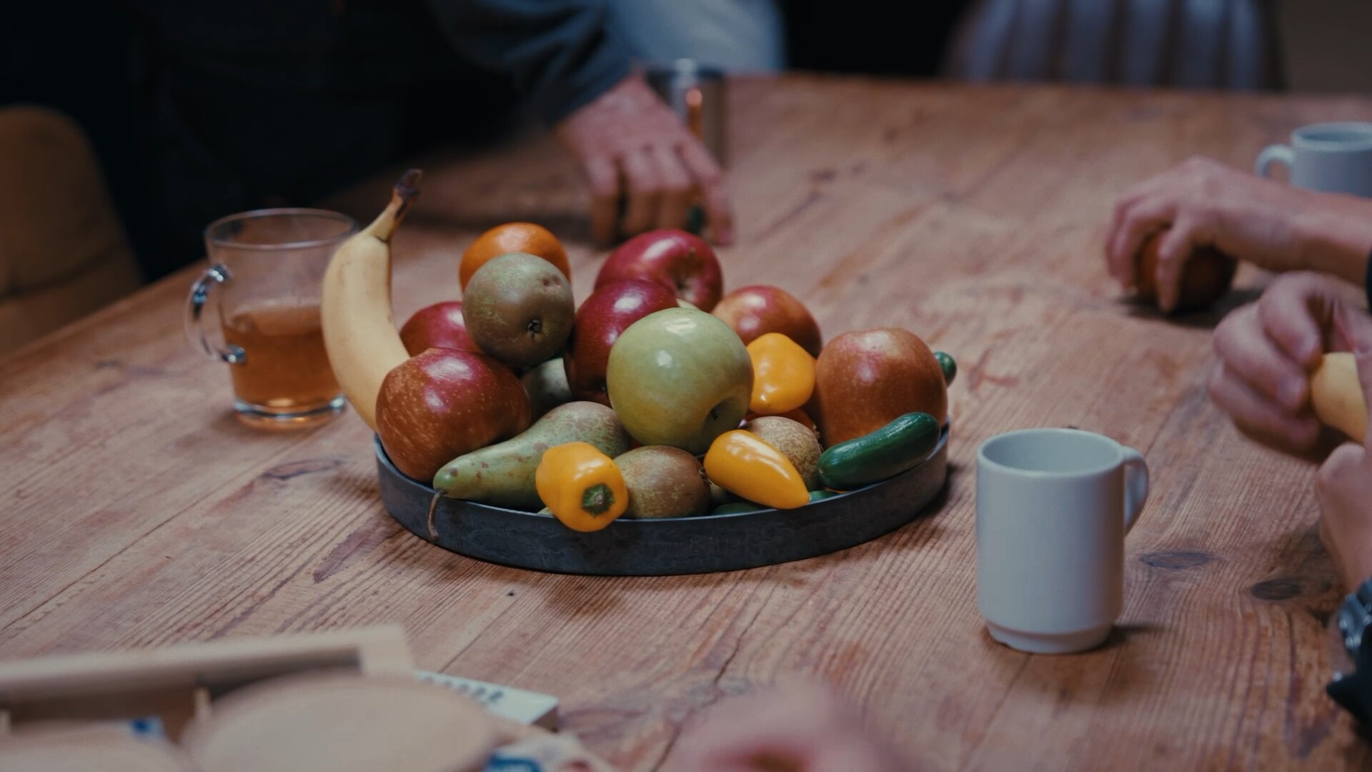 Fruitschaal, koffiemokken en een glas thee op een houten tafel tijdens een gesprek.