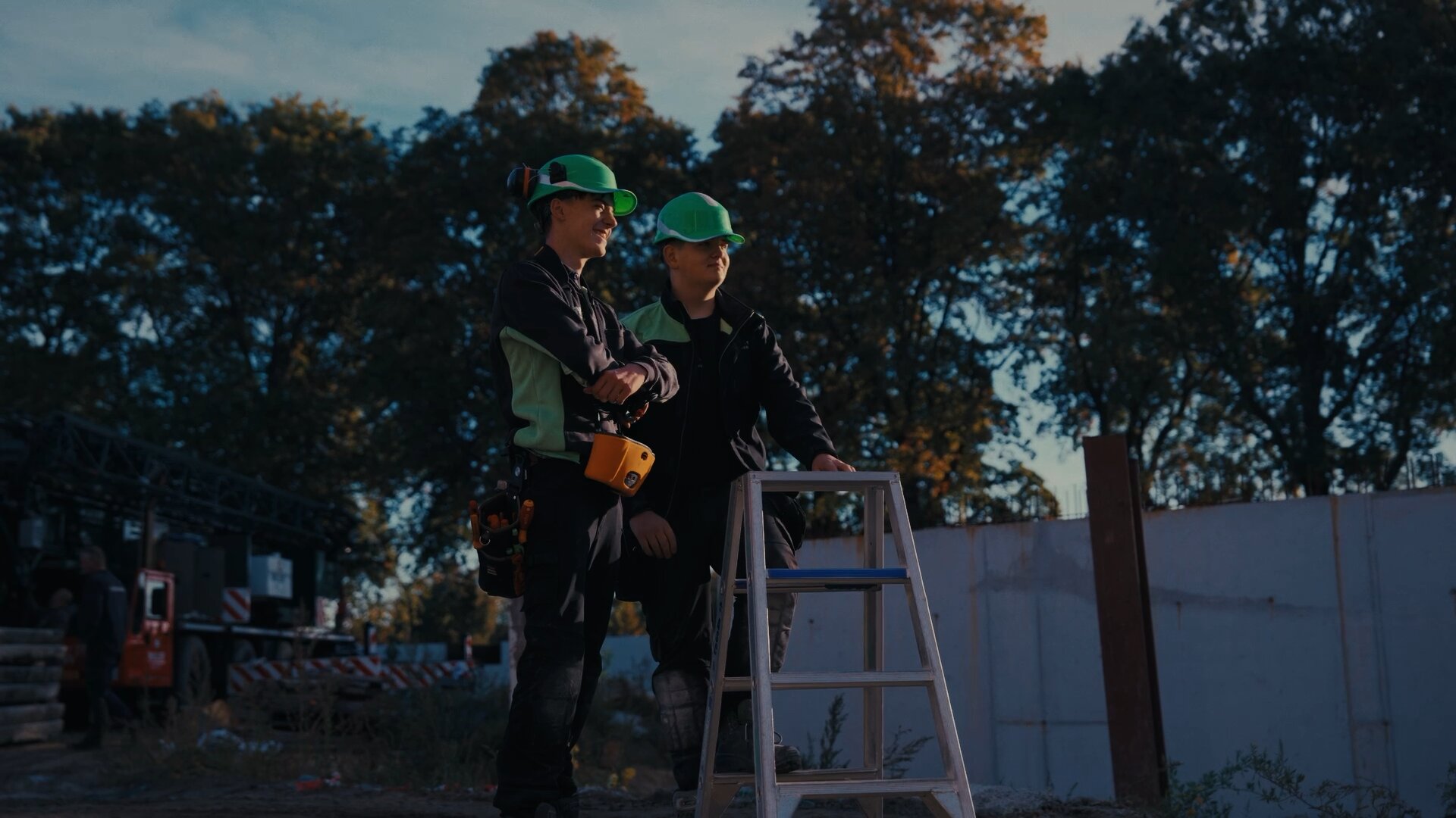 Twee monteurs met groene helmen staan lachend bij een trap op een bouwterrein.