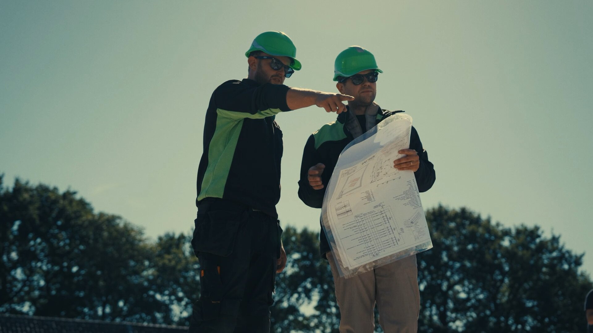 Monteur wijst iets aan terwijl een collega de bouwtekening vasthoudt, buiten op de bouw.