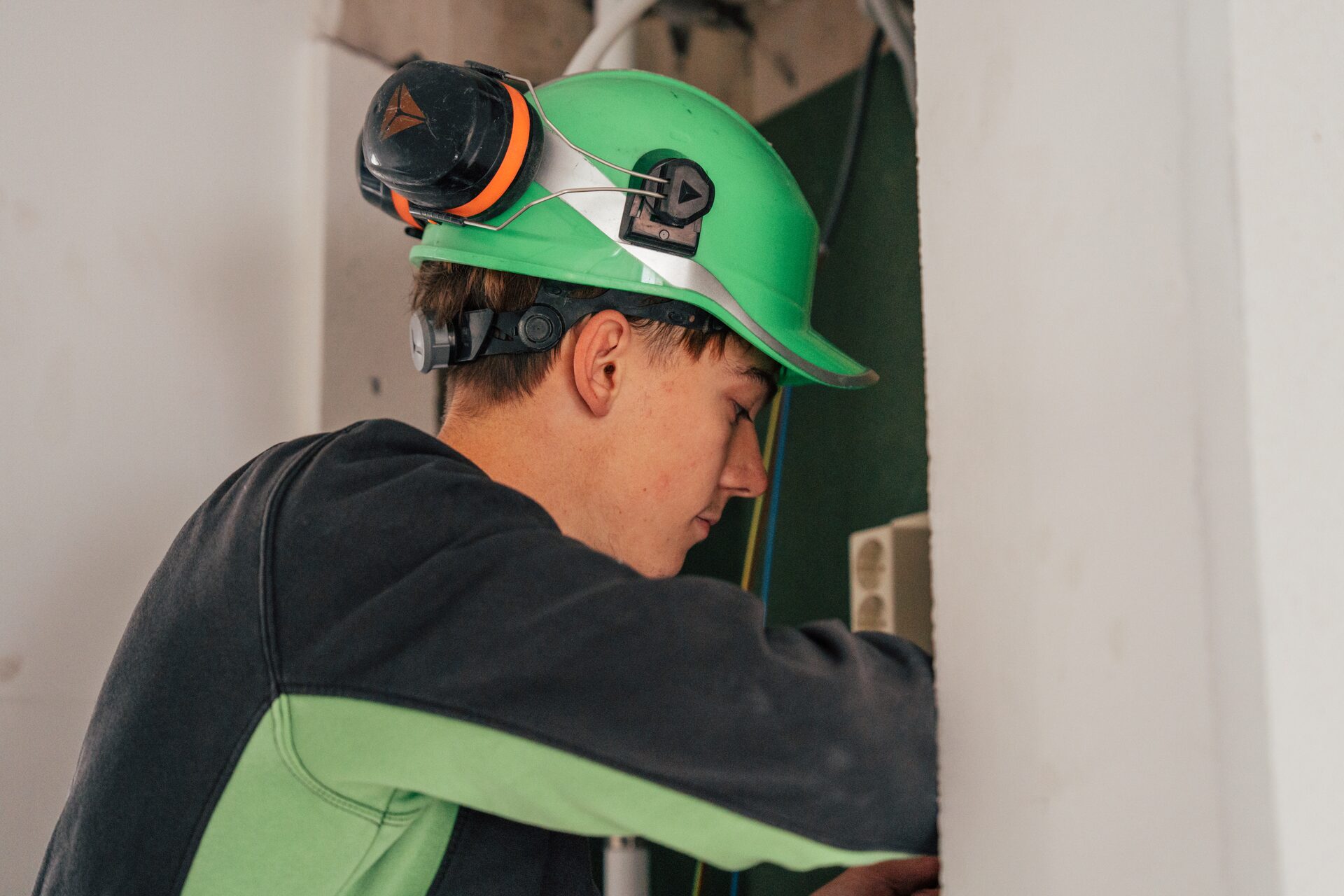 Monteur met groene helm en gehoorkappen werkt aan de bedrading in een nieuwbouwwoning.