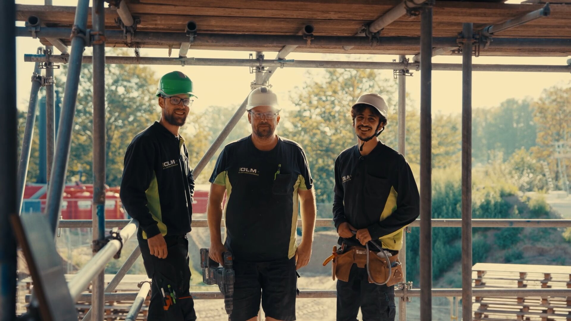 Drie monteurs van verschillende leeftijden lachen naar de camera op een bouwplaats met steigers.