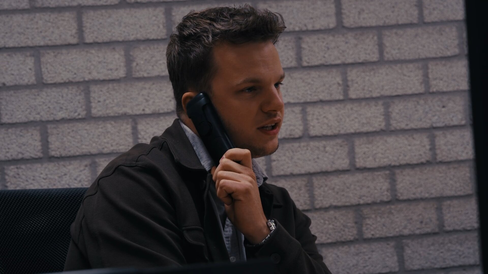Medewerker voert aan het bureau een telefoongesprek op kantoor, tegen een witte bakstenen muur.