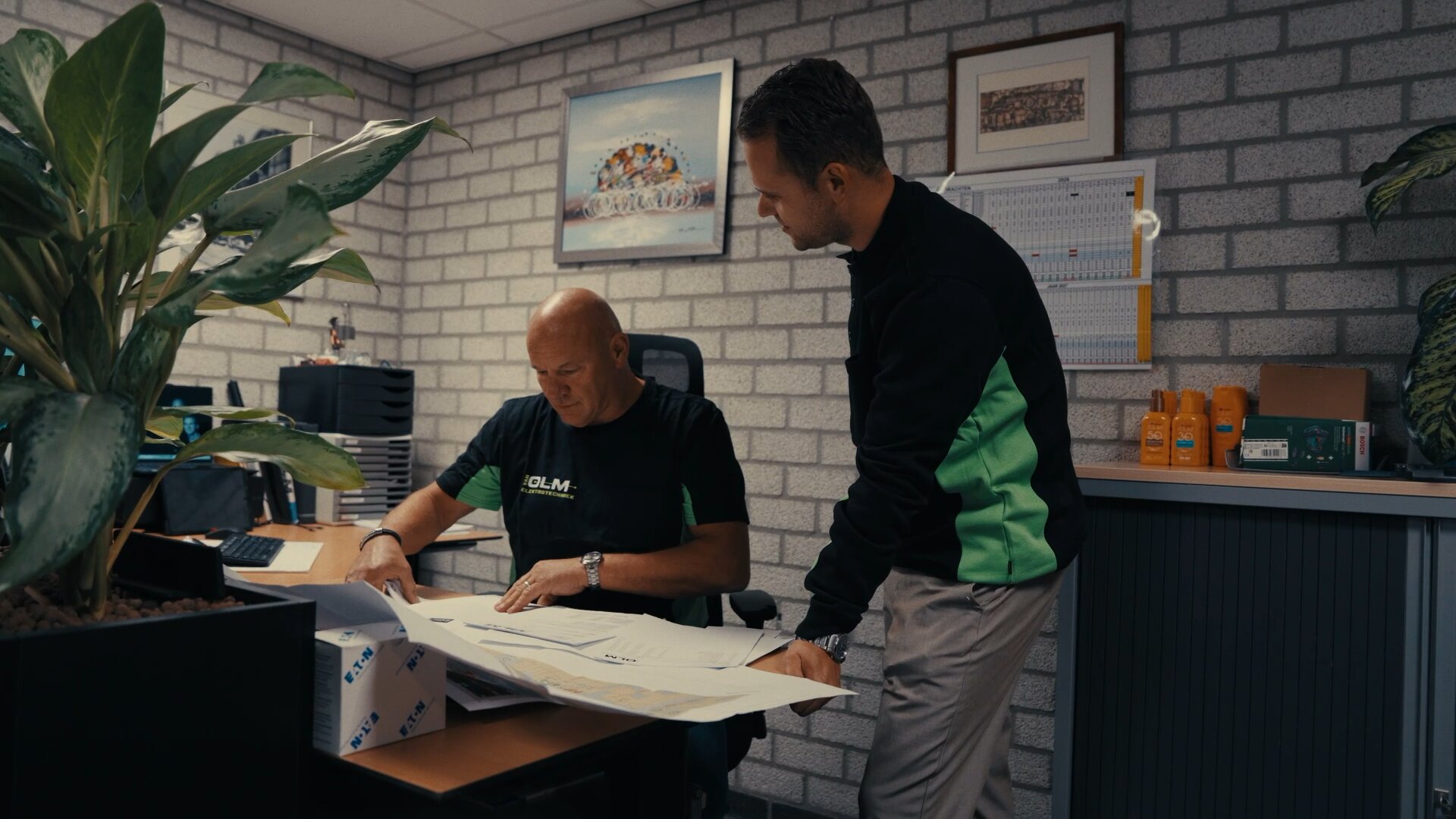 Medewerker in Van Olm-shirt rolt bouwtekeningen uit op het bureau, een collega kijkt mee.