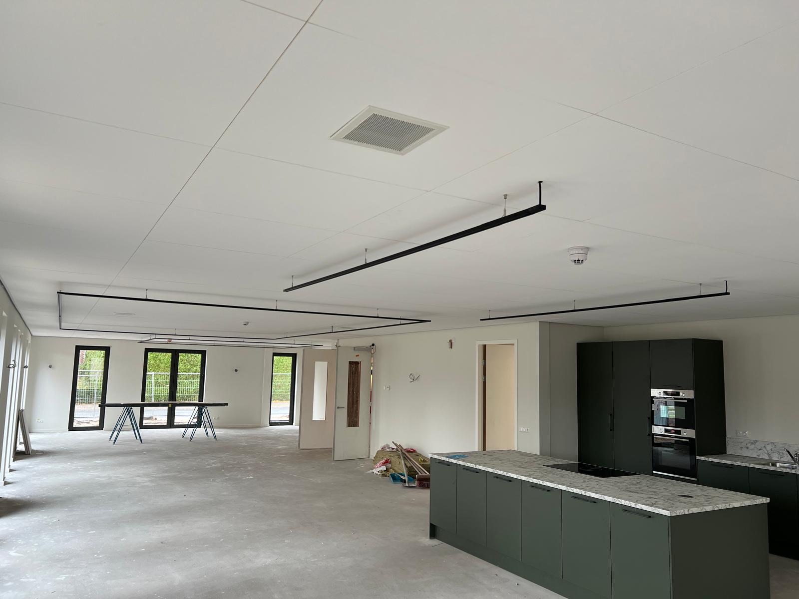 Zwarte lichtrails aan een wit plafond boven een open ruimte in afbouw, met een keukenblok rechts.