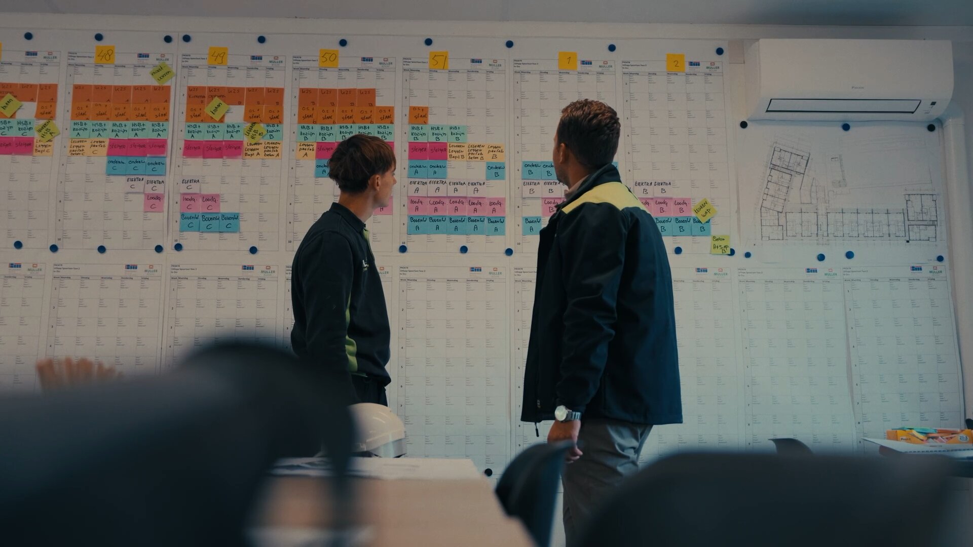 Twee collega's bekijken een wandplanning vol gekleurde memobriefjes in de bouwkeet.